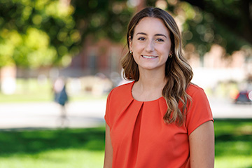 A woman wearing a red blouse smiles at the camera while standing outdoors on a university campus, photographed for a professional portrait.