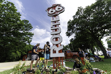 A group of children dig and plant flowers around a large Route 66 sign in a landscaped garden. Trees and a vintage red fire truck are visible in the background.