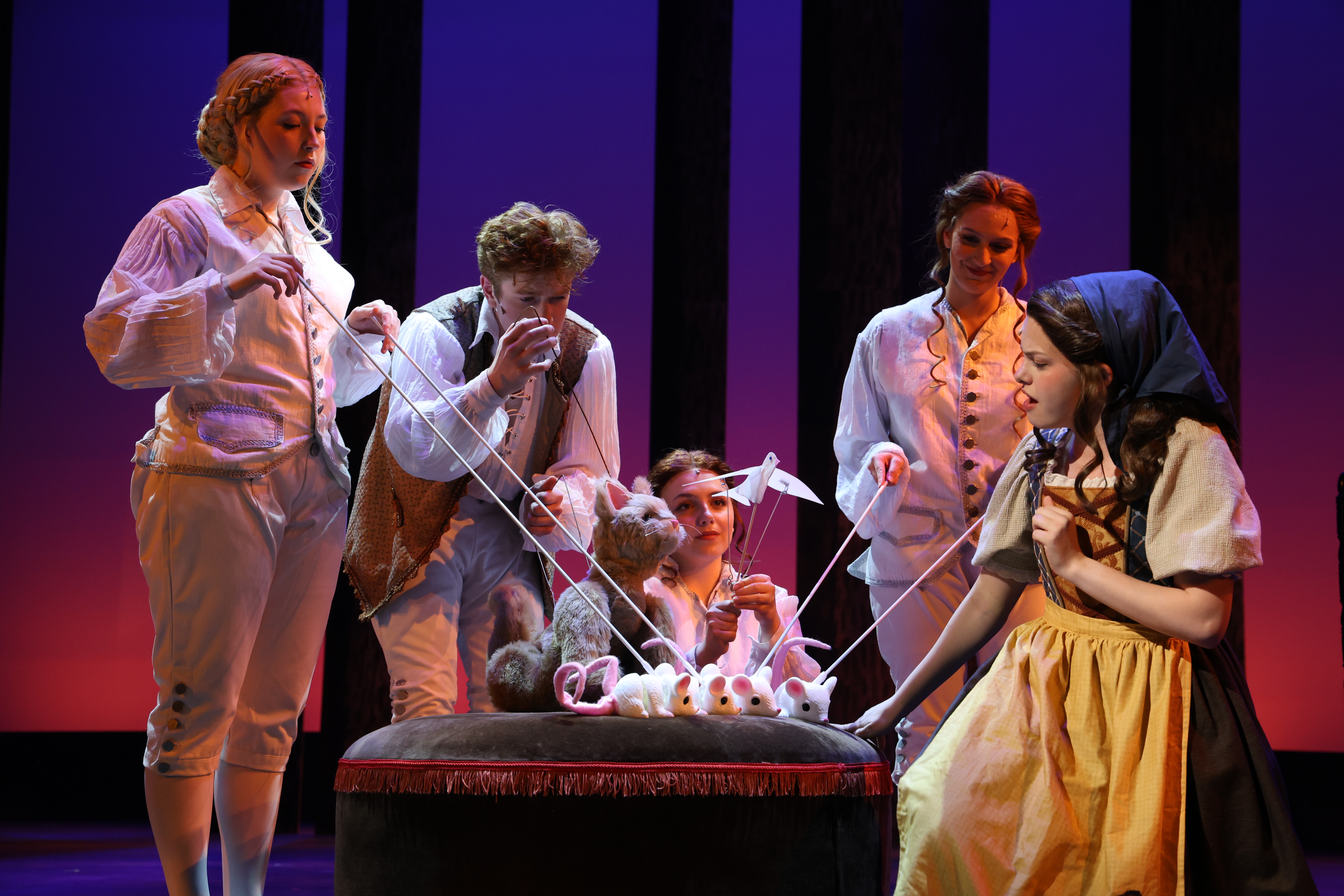 A stage scene from a theatrical production shows performers in period costumes gathered around a raised table, manipulating string puppets shaped like mice beside a seated central character under colorful theatrical lighting.