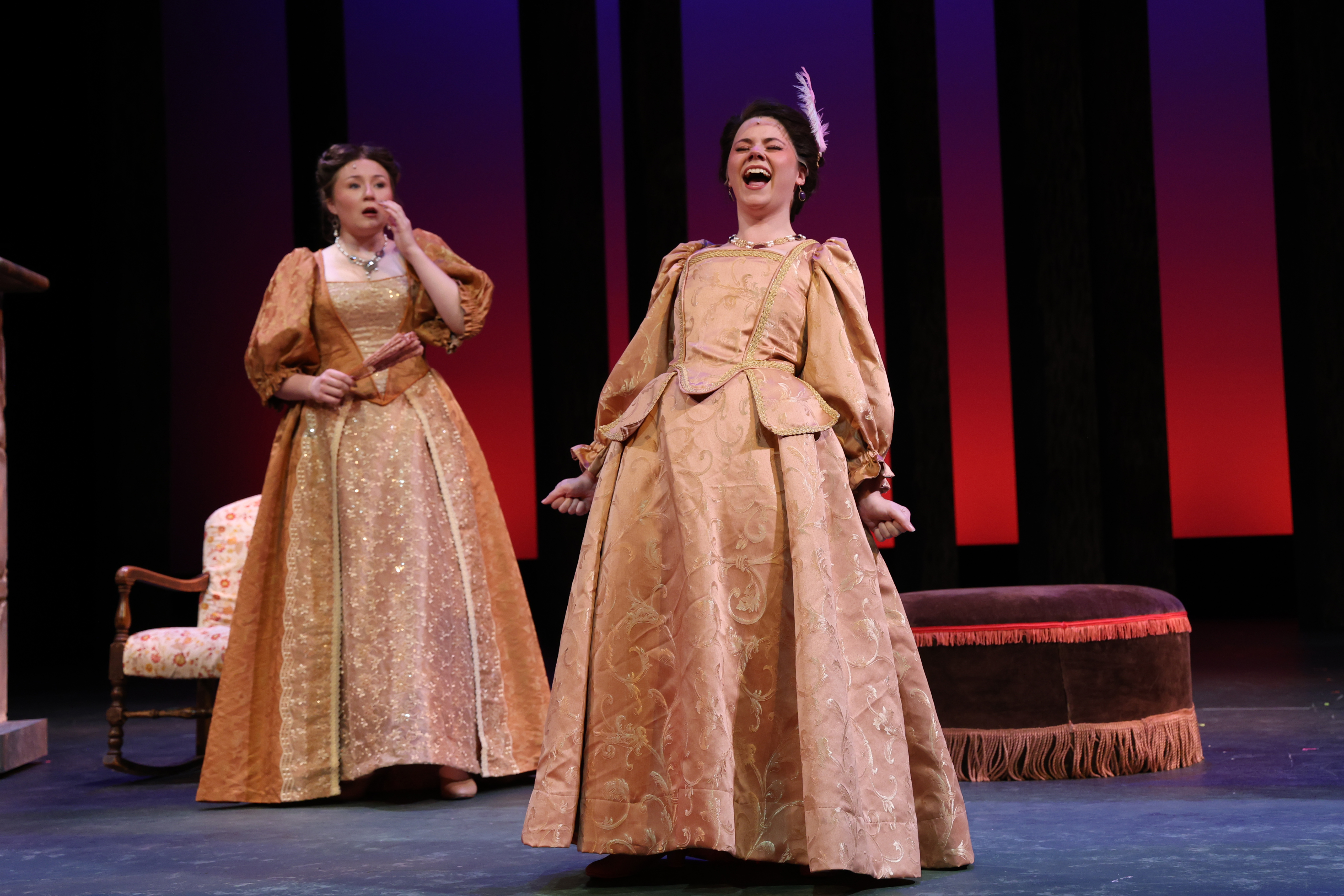 A stage scene from a theatrical production shows two performers in ornate period costumes in the foreground—one reacting dramatically while another performs—set against a striped backdrop with stage furniture and theatrical lighting.