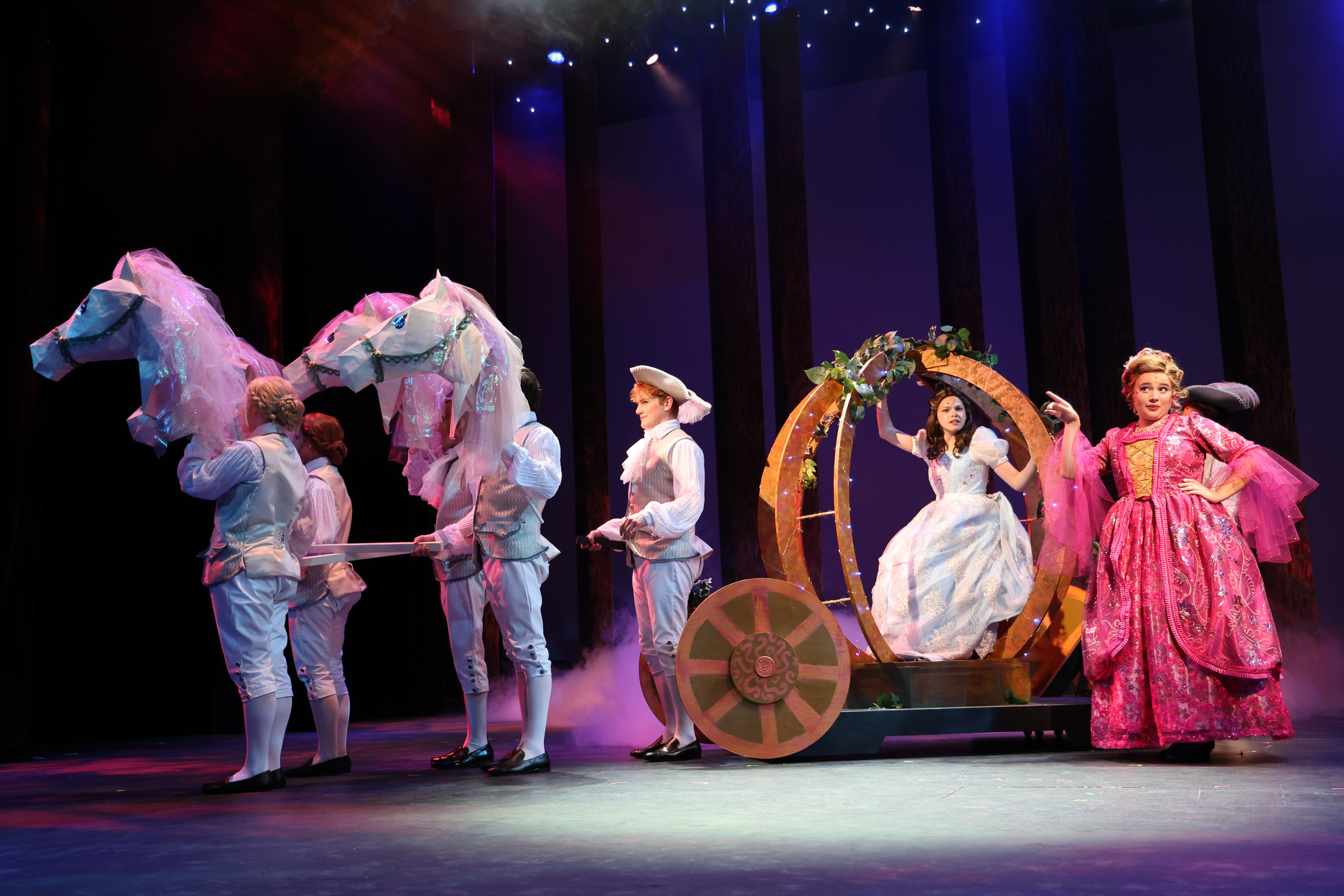 A stage scene from a theatrical production shows performers in period costumes escorting a character seated in a decorative carriage while others hold stylized horse figures, depicting a magical transformation moment under colorful stage lighting.