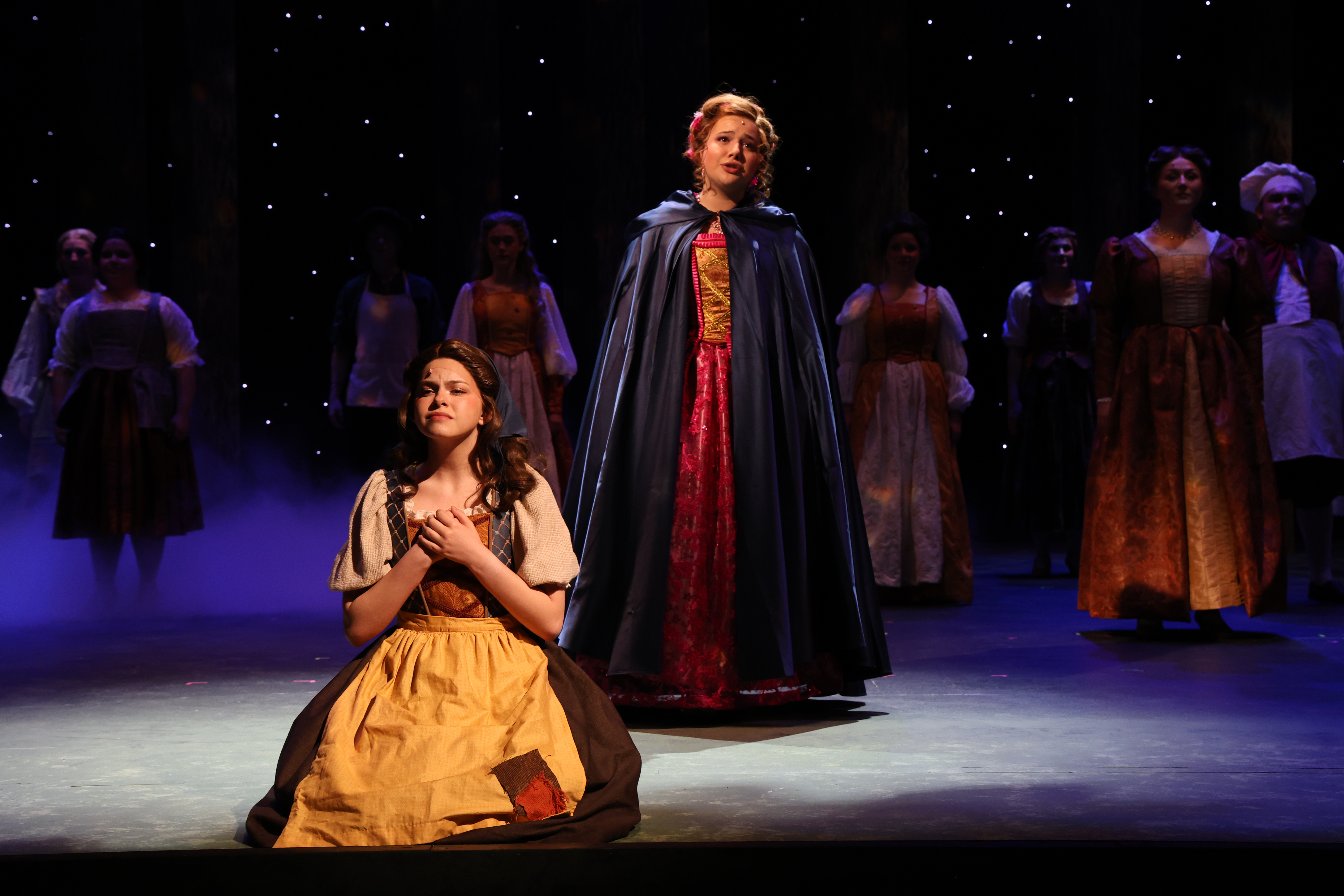 A stage scene from a theatrical production shows one performer kneeling in the foreground while another stands and sings behind them, with an ensemble in period costumes arranged across a dimly lit stage under a star‑like backdrop.