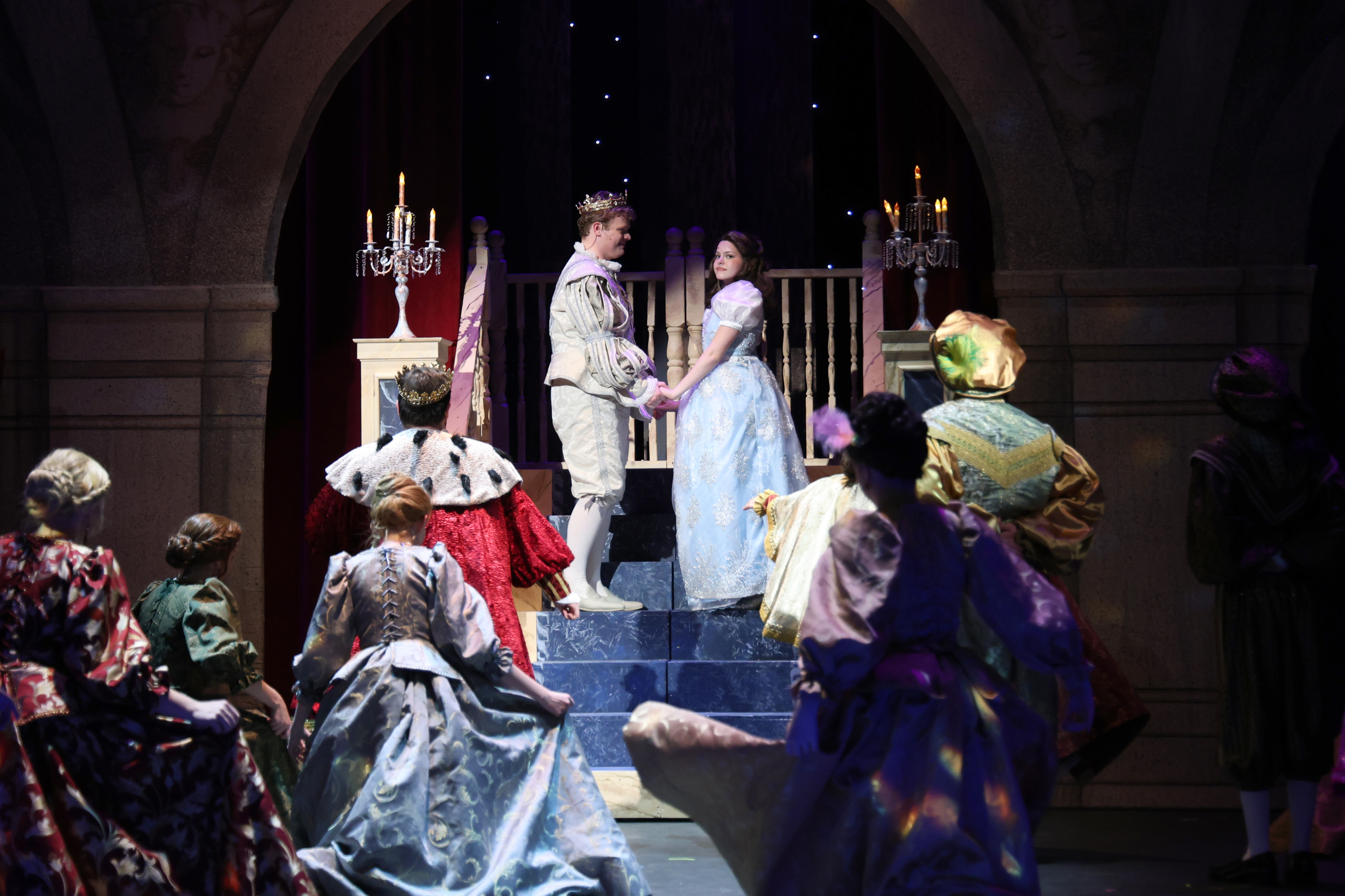 A final stage tableau from a theatrical production shows two performers in royal costumes holding hands atop steps beneath an arched set, surrounded by an ensemble in formal period attire and candlelit décor to depict a celebratory court scene.