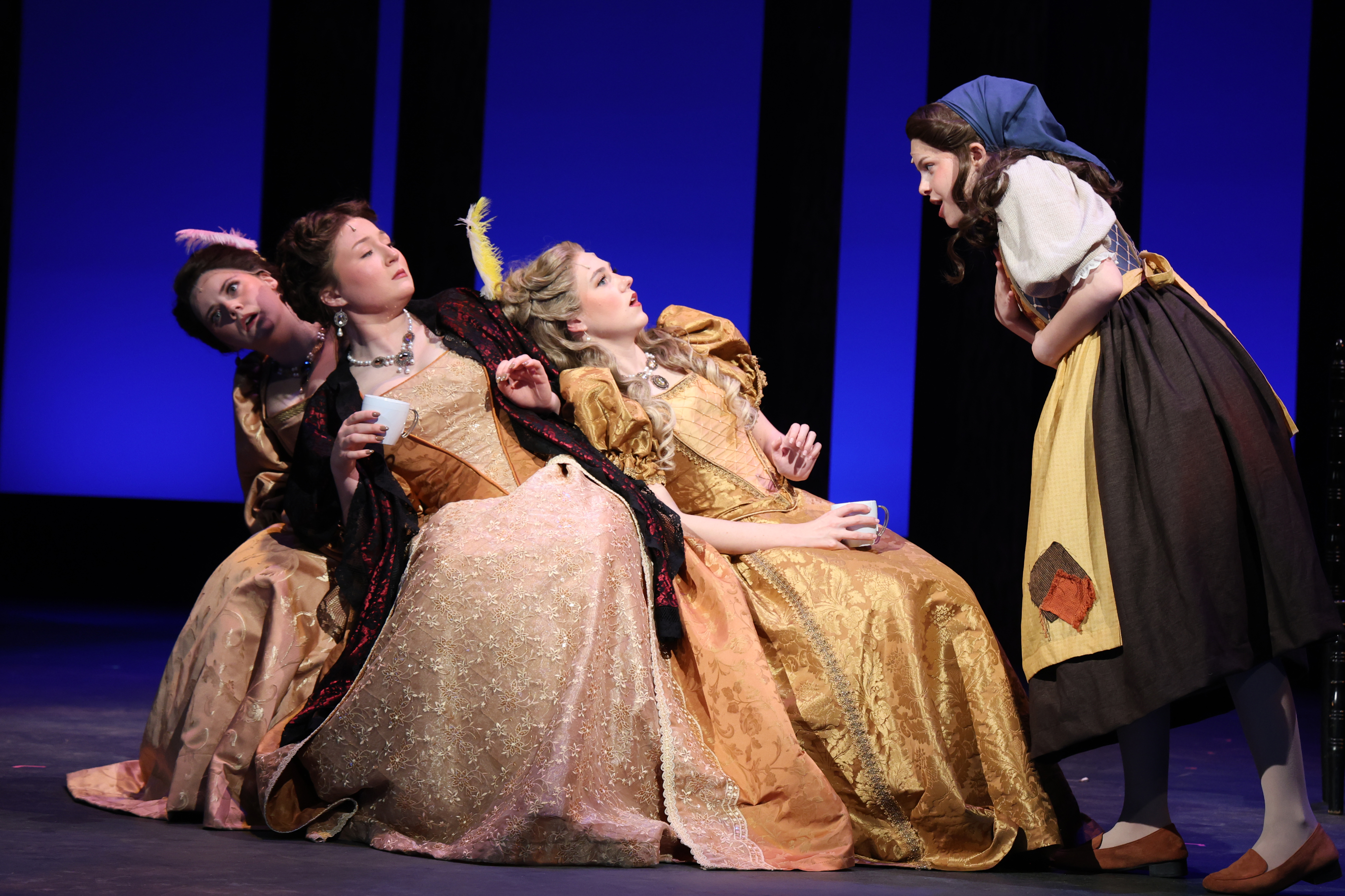 A stage scene from a theatrical production shows several performers in ornate period costumes leaning together on a bench while another performer in simpler clothing stands to the side addressing them, set against a blue‑lit backdrop with vertical panels.