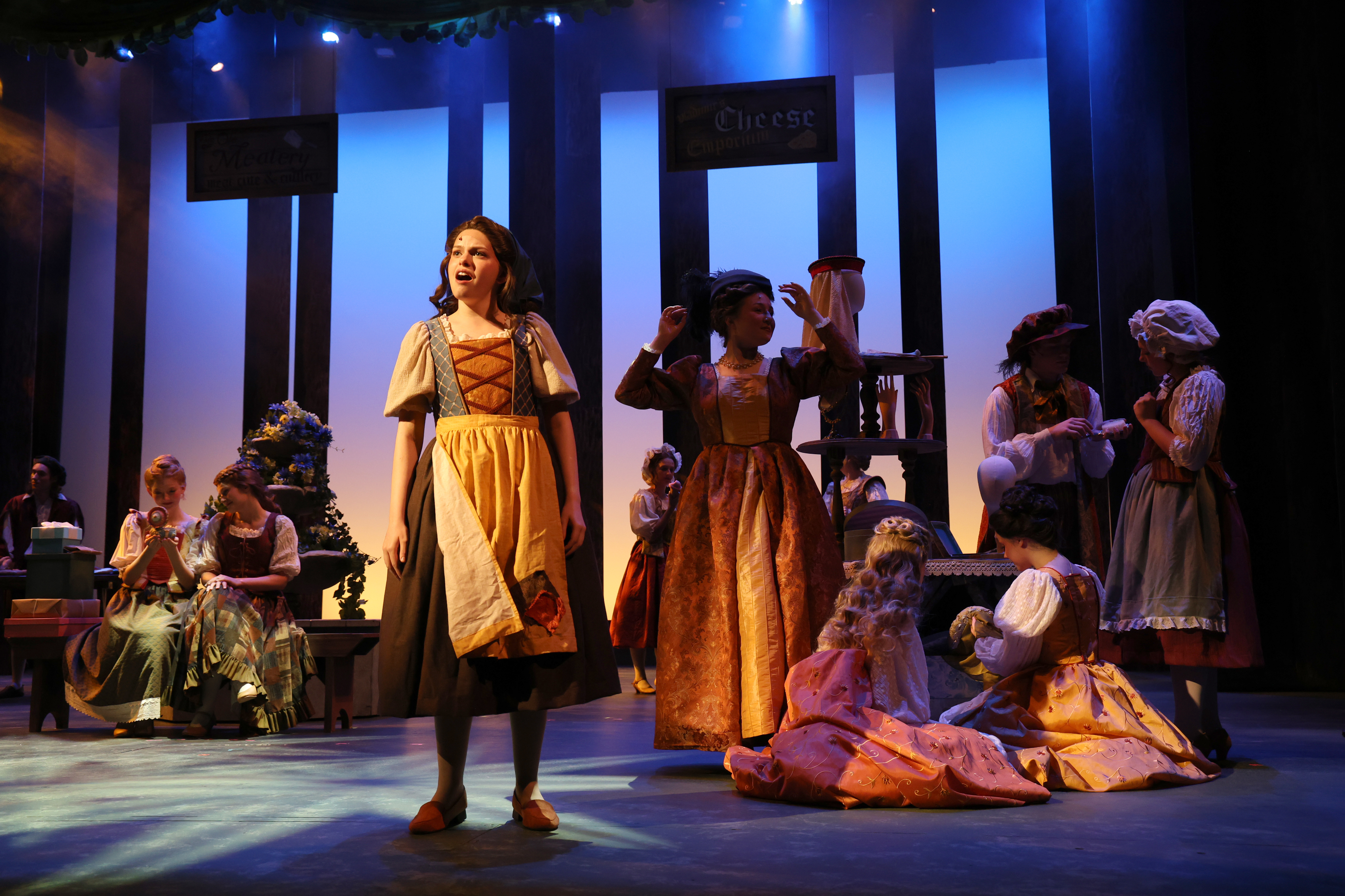 A stage scene from a theatrical production shows a central performer in period costume standing under bright stage lighting while an ensemble of performers in similar historical attire gather and act around them on a stylized village set.