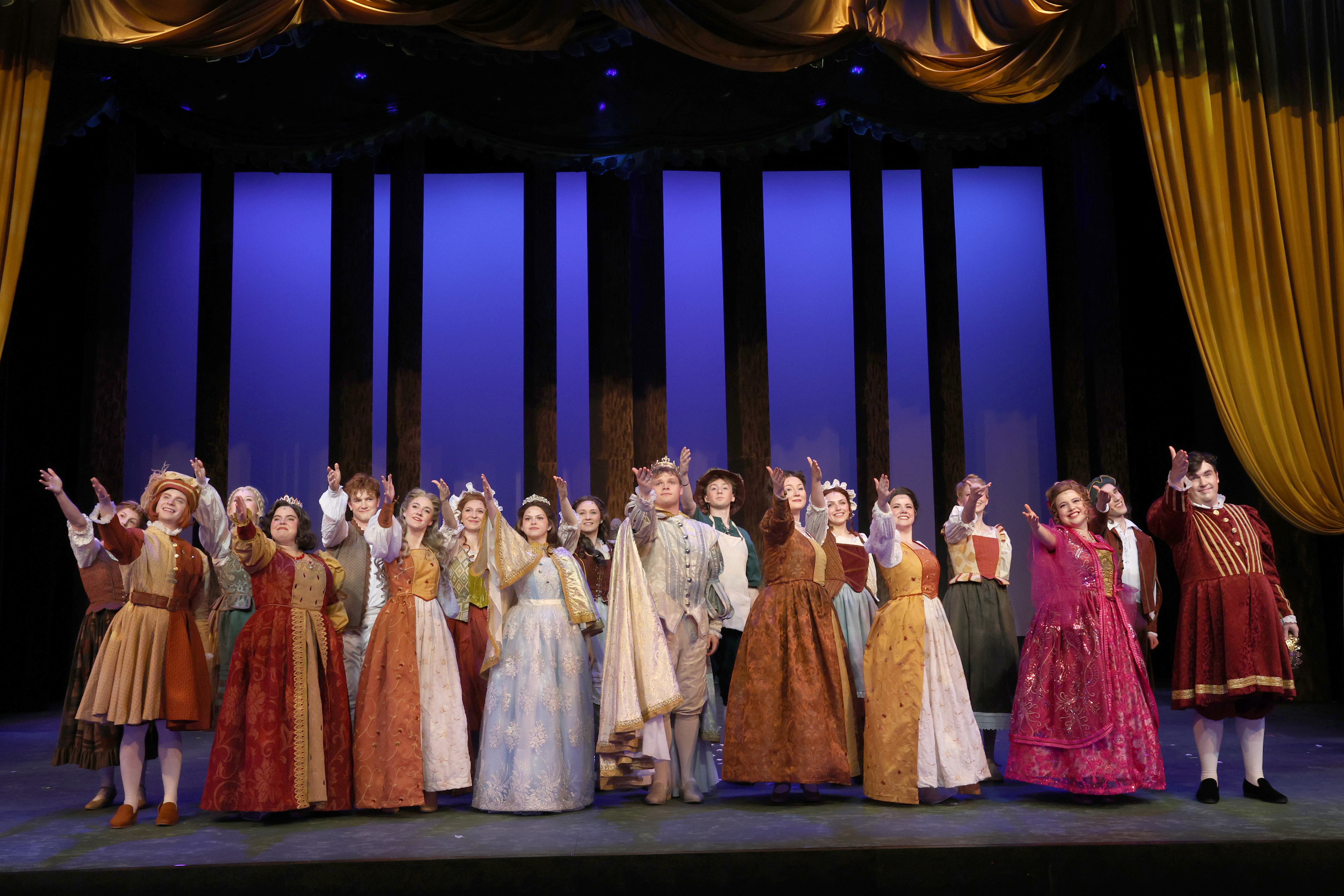 A full cast of performers in colorful period costumes stands across a theater stage with arms raised during the final curtain call, framed by tall vertical panels and warm stage lighting to conclude the theatrical production.