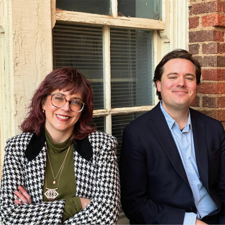 Two people sit side by side in front of an old multi‑pane window and brick wall. The person on the left wears a houndstooth jacket over a green top with layered necklaces, while the person on the right wears a dark blazer over a light blue shirt.