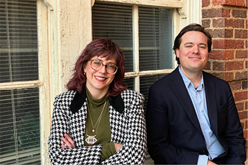 Two people sit side by side in front of an old multi‑pane window and brick wall. The person on the left wears a houndstooth jacket over a green top with layered necklaces, while the person on the right wears a dark blazer over a light blue shirt.