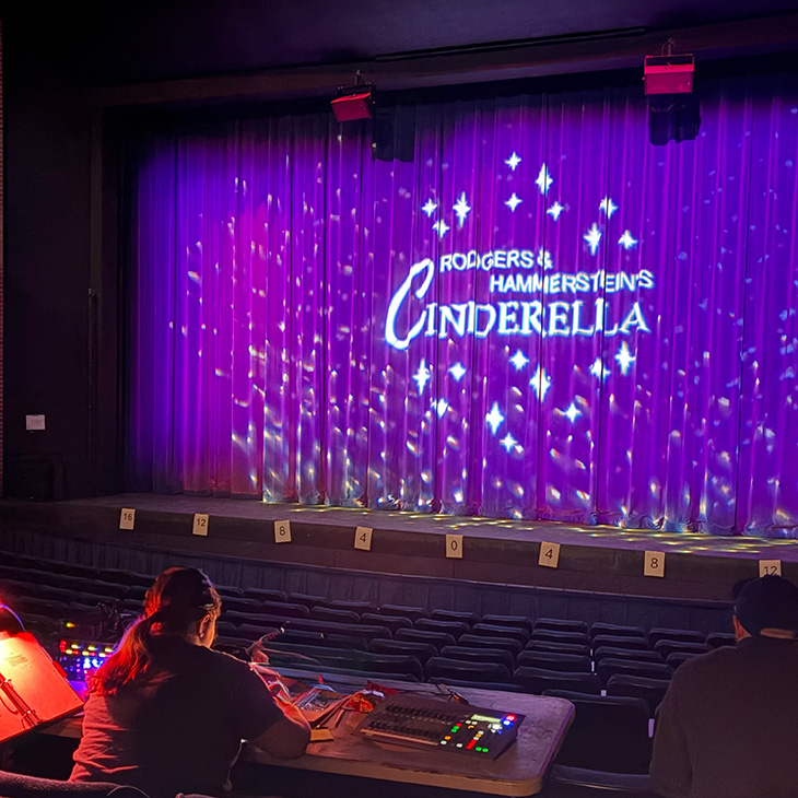 A theater stage is lit with purple lighting and a backdrop reading “Rodgers & Hammerstein’s Cinderella,” while technical staff sit at a control table in the audience area, indicating pre-show or production setup.