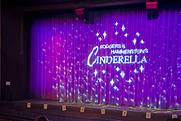 A theater stage is lit with purple lighting and a backdrop reading “Rodgers & Hammerstein’s Cinderella,” while technical staff sit at a control table in the audience area, indicating pre-show or production setup.