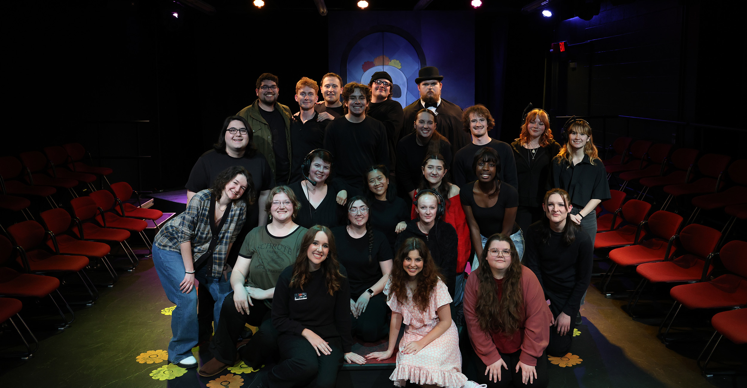 Group photo of a theatre cast and crew posed together on a stage, with rows of red audience chairs and stage lighting visible in the background.