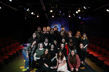 Group photo of a theatre cast and crew posed together on a stage, with rows of red audience chairs and stage lighting visible in the background.