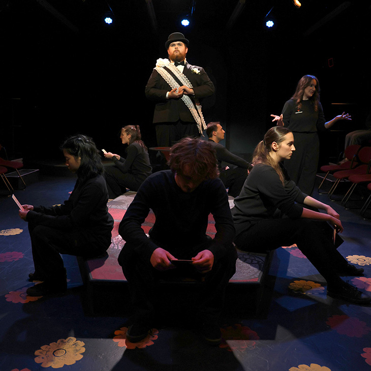 Theatrical scene from a stage production shows an ensemble of performers in black clothing arranged in a circular composition, with one central figure standing elevated while others sit or gesture under dramatic stage lighting.