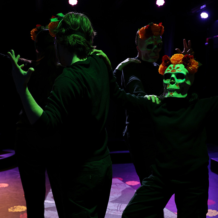 Stage performance scene shows several performers in black costumes moving together under dramatic green and purple lighting, with one wearing a decorative skull mask adorned with flowers, creating a ritualistic, expressive tableau.