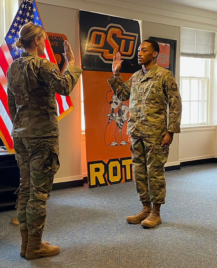 Two individuals in military uniforms stand facing each other with raised right hands during a swearing‑in or oath ceremony, with an American flag and OSU ROTC banner displayed behind them indoors.