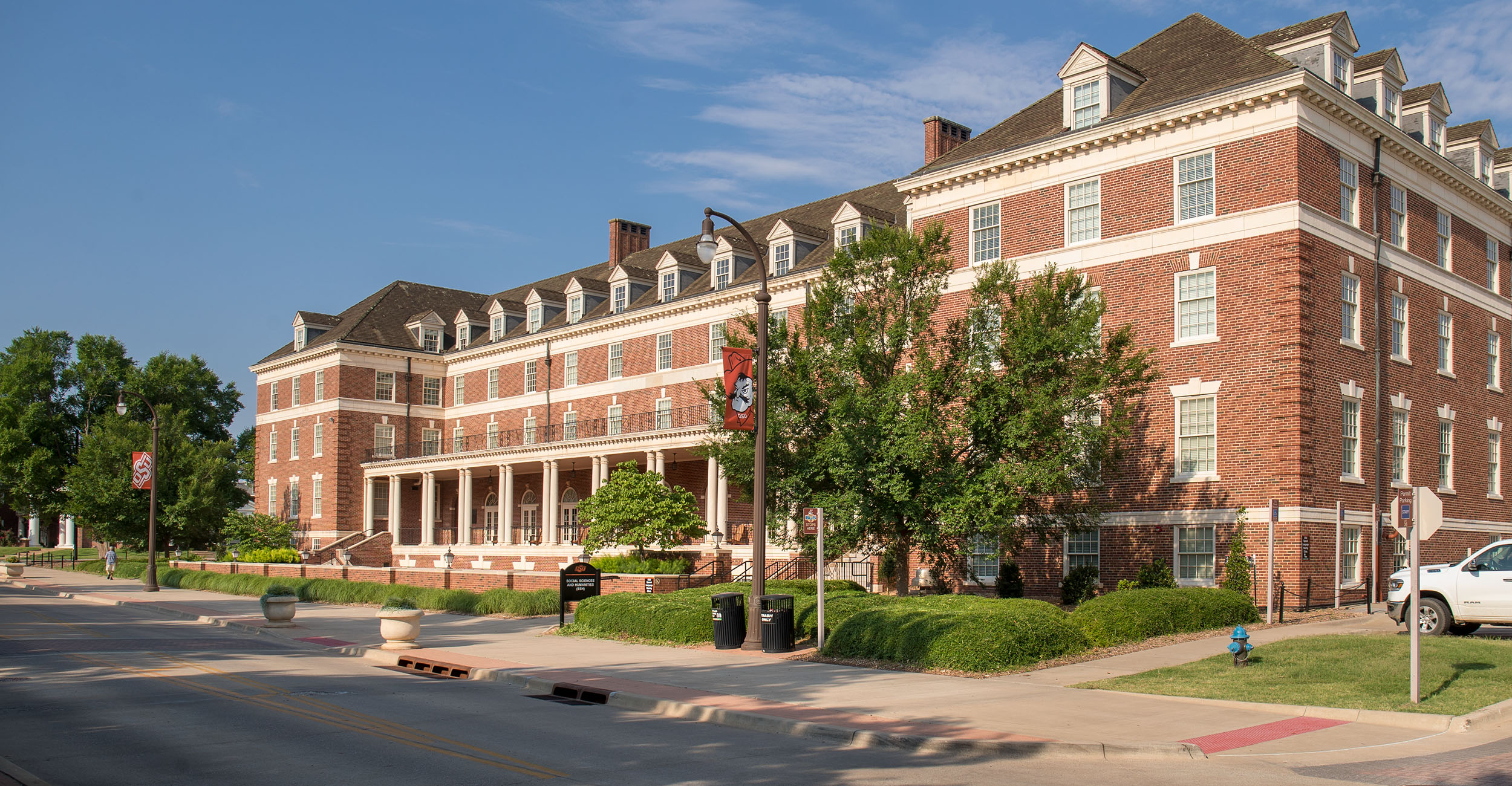 A large red-brick building with white trim and multiple dormer windows, featuring a columned front porch and landscaped greenery along the sidewalk under a clear blue sky.