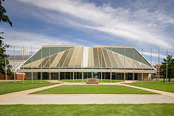 Front view of Oklahoma State University’s Noble Research Center, showing the building’s distinctive sloped roof, flagpoles, landscaped lawn, and walkways under a partly cloudy sky.