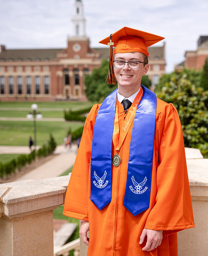 A graduate wearing an orange cap and gown with a blue military service stole and medallion stands on campus in front of a historic academic building, posed for a formal graduation portrait.