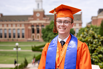 A graduate wearing an orange cap and gown with a blue military service stole and medallion stands on campus in front of a historic academic building, posed for a formal graduation portrait.
