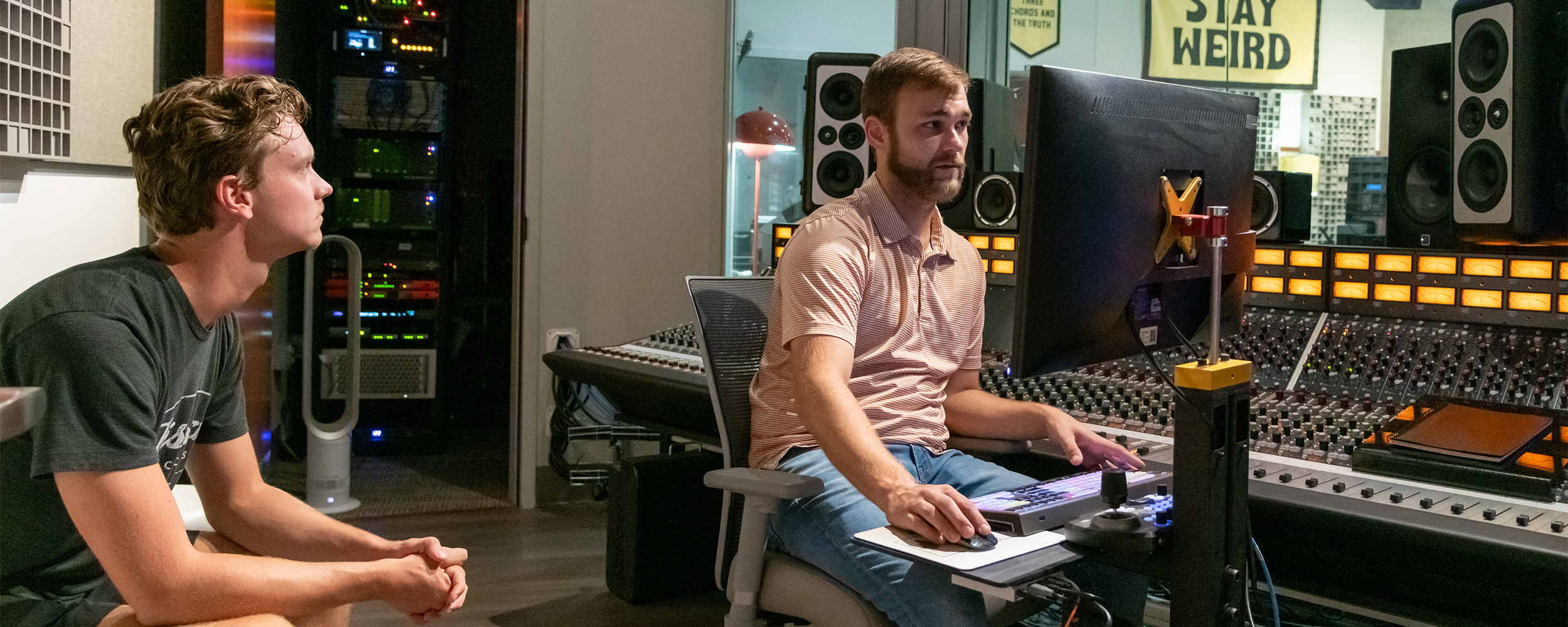 Mitchell Crawford works at a mixing console alongside a student in an audio recording studio.