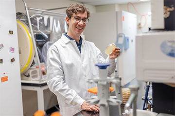 Dr. Paul Kirchberger smiles while holding a small laboratory sample container surrounded by scientific equipment.