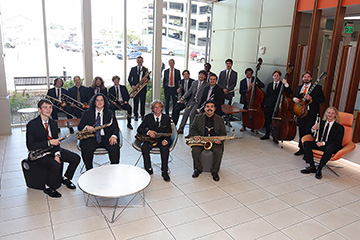 Members of the Oklahoma State University Jazz Orchestra pose with saxophones, trombones, trumpets, upright bass and guitar in a bright lobby.