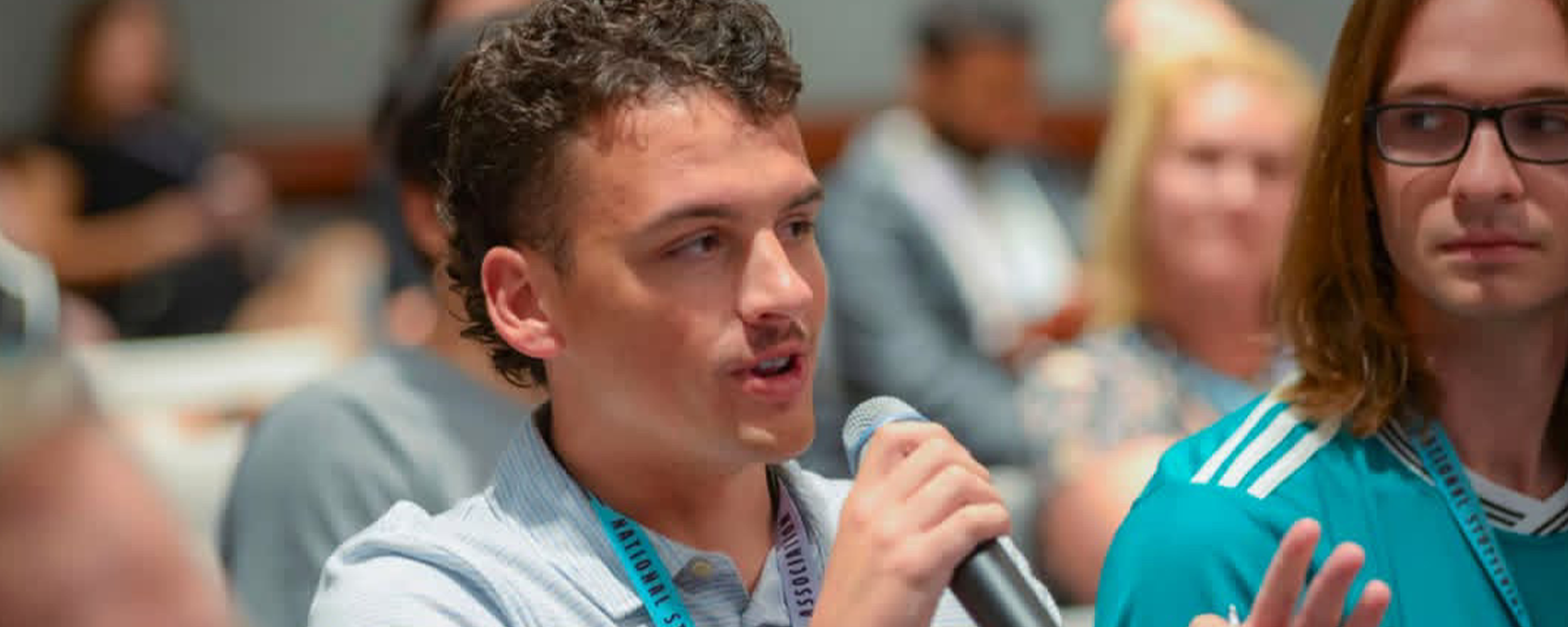 Zachary Ormsby speaks into a microphone during a National Stuttering Association event as fellow attendees listen in the background.