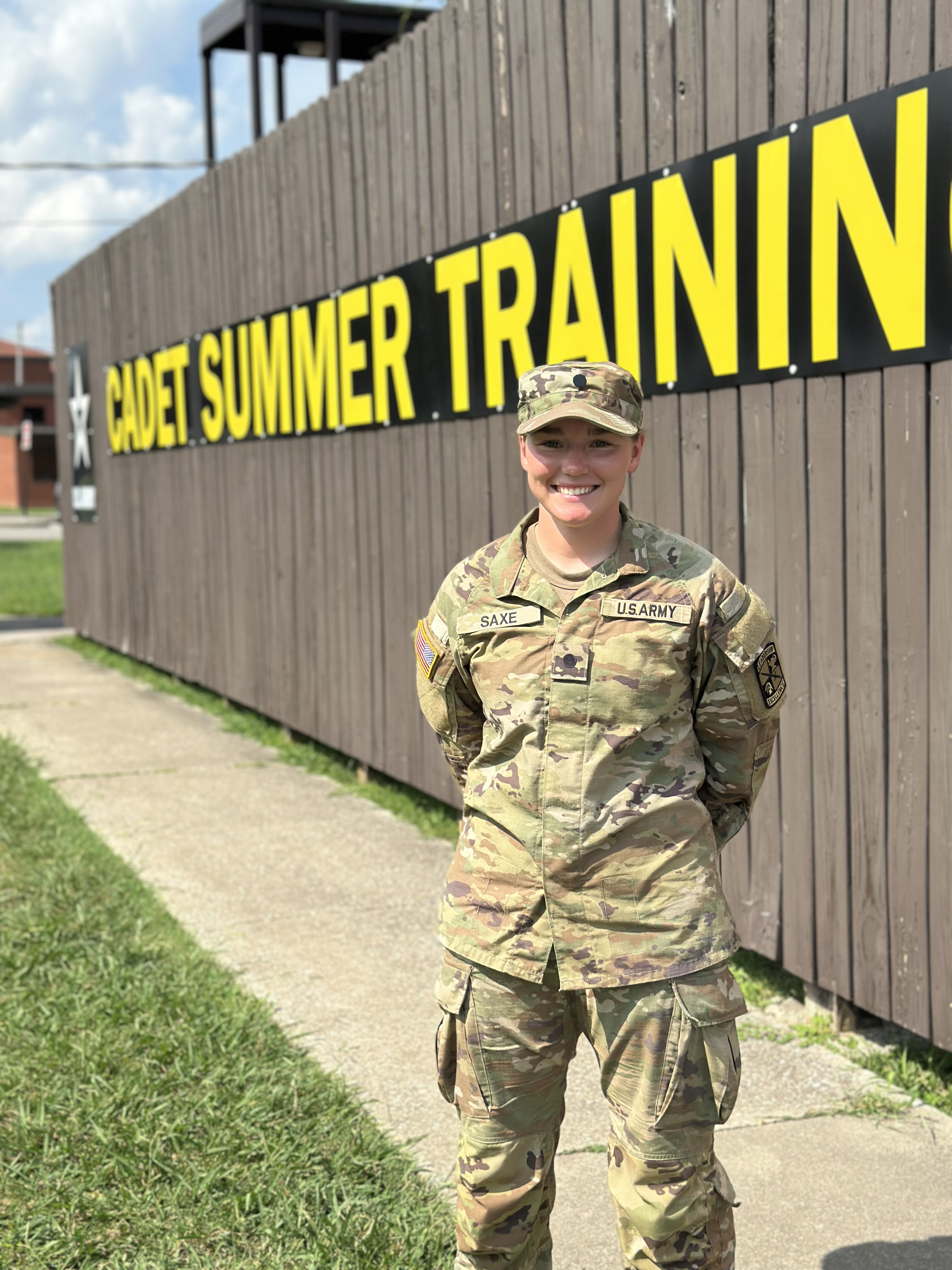 Emma Saxe stands in uniform in front of a Cadet Summer Training sign at Fort Knox.