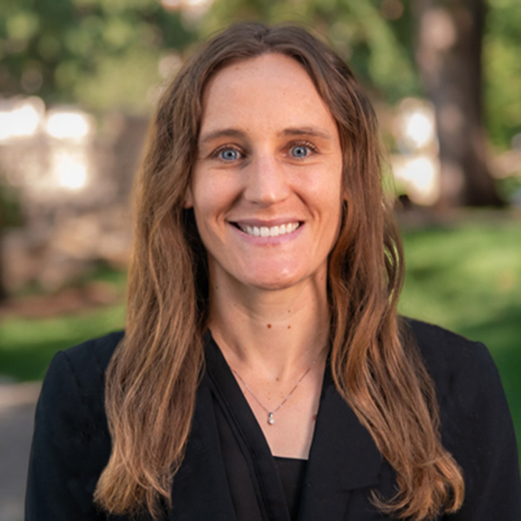 Person with long wavy hair wearing a black blazer and a delicate necklace, standing outdoors with green grass and trees in the background.