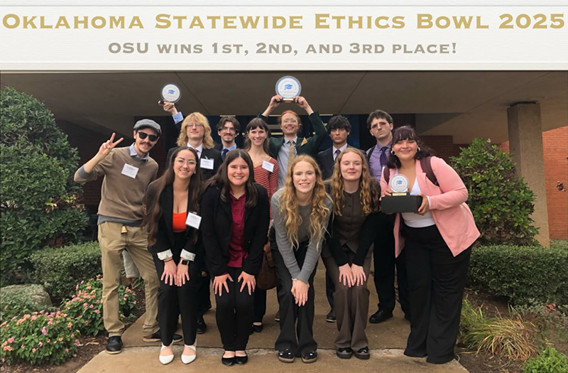 A group of people posing outdoors in front of a banner that reads “Oklahoma Statewide Ethics Bowl 2025 – OSU wins 1st, 2nd, and 3rd place!” Several individuals are holding award plaques, and the group is dressed in business and semi-formal attire.
