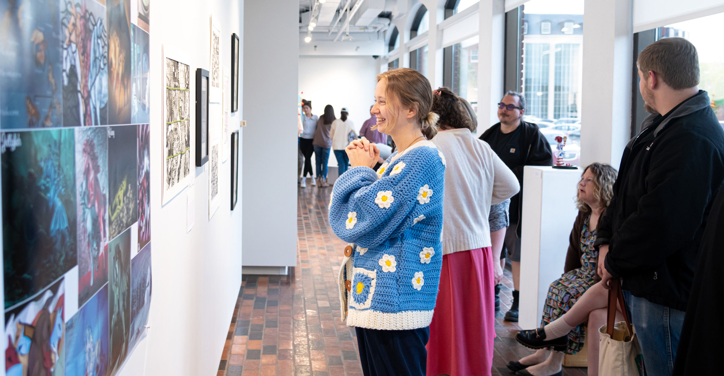 A young blonde woman wearing a blue sweater smiles and clasps her hands as she looks at art on the wall of the Gardiner Gallery.