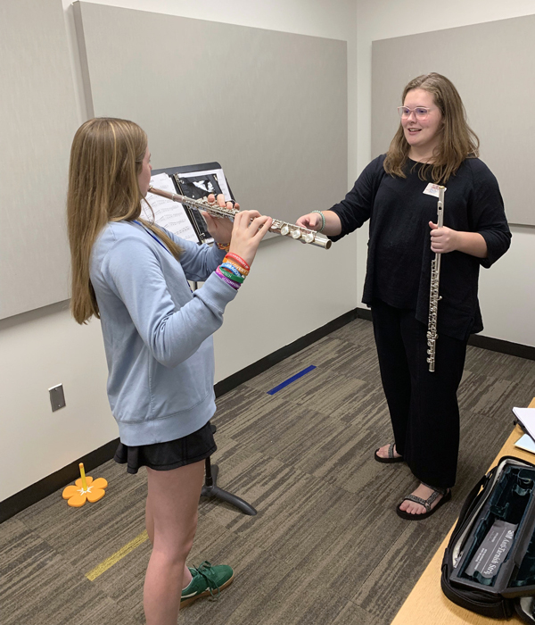 A young woman stands by a music stand holding a flute and instructs a high school student also holding a flute.