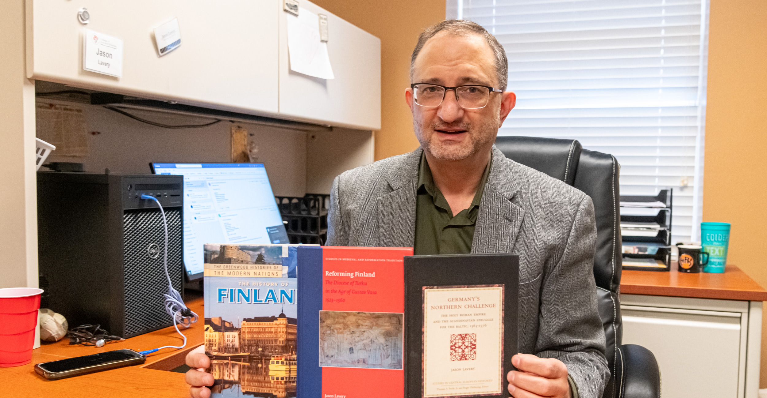 Dr. Jason Lavery sits at his desk and displays his three published books.
