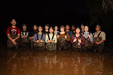 A group of students and an instructor wearing chest waders and headlamps stand together in shallow water at night during a hands-on field research activity.