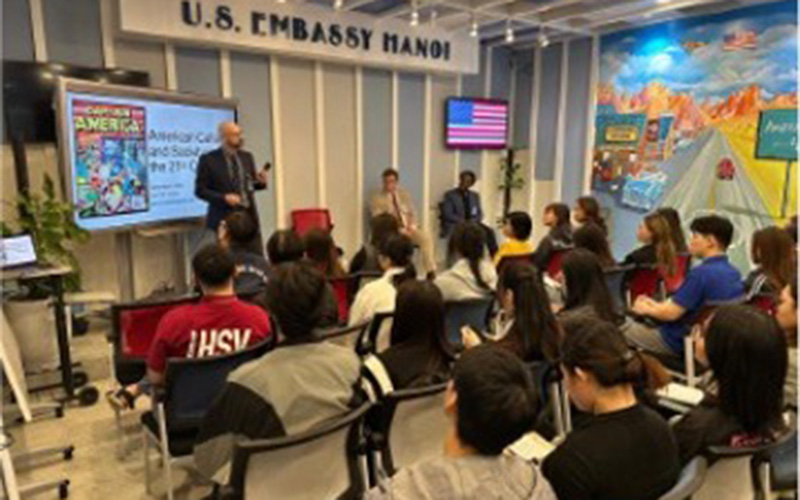 An audience of students listens to a speaker giving an American Studies presentation inside the U.S. Embassy in Hanoi, with educational posters and a U.S. flag displayed at the front of the room.