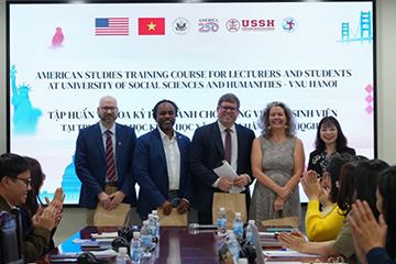 A group of presenters stands on a stage in front of a projected banner for an American Studies training course at the University of Social Sciences and Humanities in Hanoi, indicating an academic international collaboration event.
