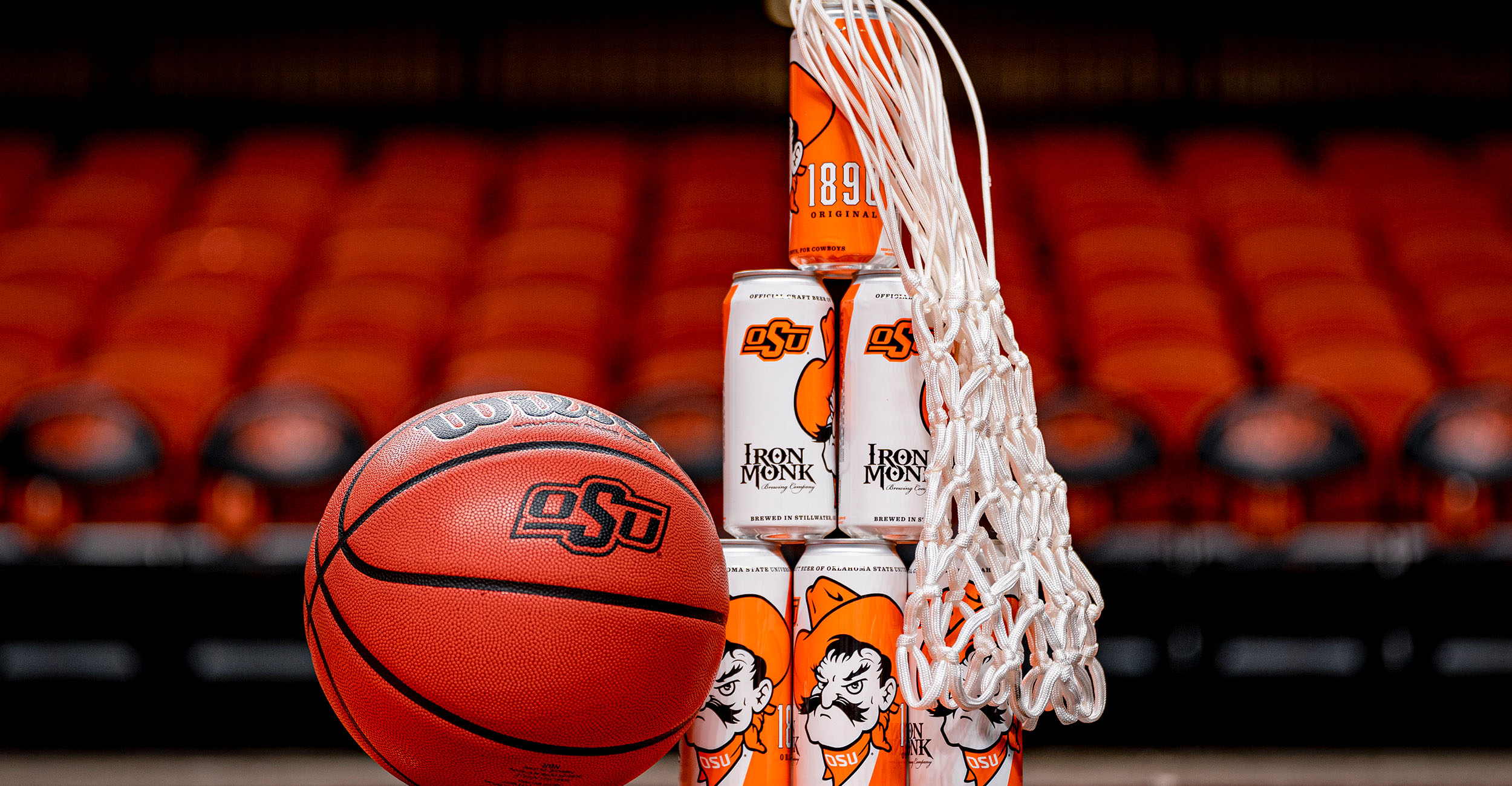 1890 Original from Iron Monk is the official craft brew of Oklahoma State, and it is pictured in Gallagher-Iba Arena next to a basketball and a net.