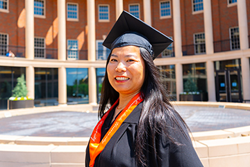 A person wearing a graduation cap and academic attire with an orange stole stands in an outdoor campus courtyard with columns and a brick building in the background, indicating a graduation portrait setting.