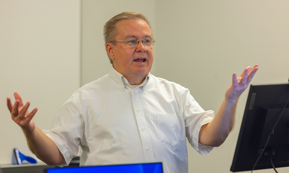 HTM faculty member Steven West teaching his class with his hands raised in the air while wearing a white shirt.