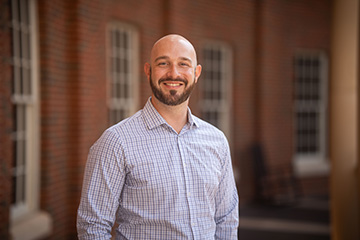 A person stands outdoors in front of a brick building, smiling at the camera in a professional headshot.
