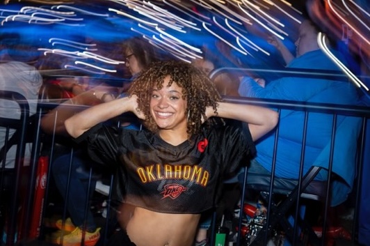A young woman with curly, brown hair smiles as she steps onto the court at Paycom Center. Lights shine around her.