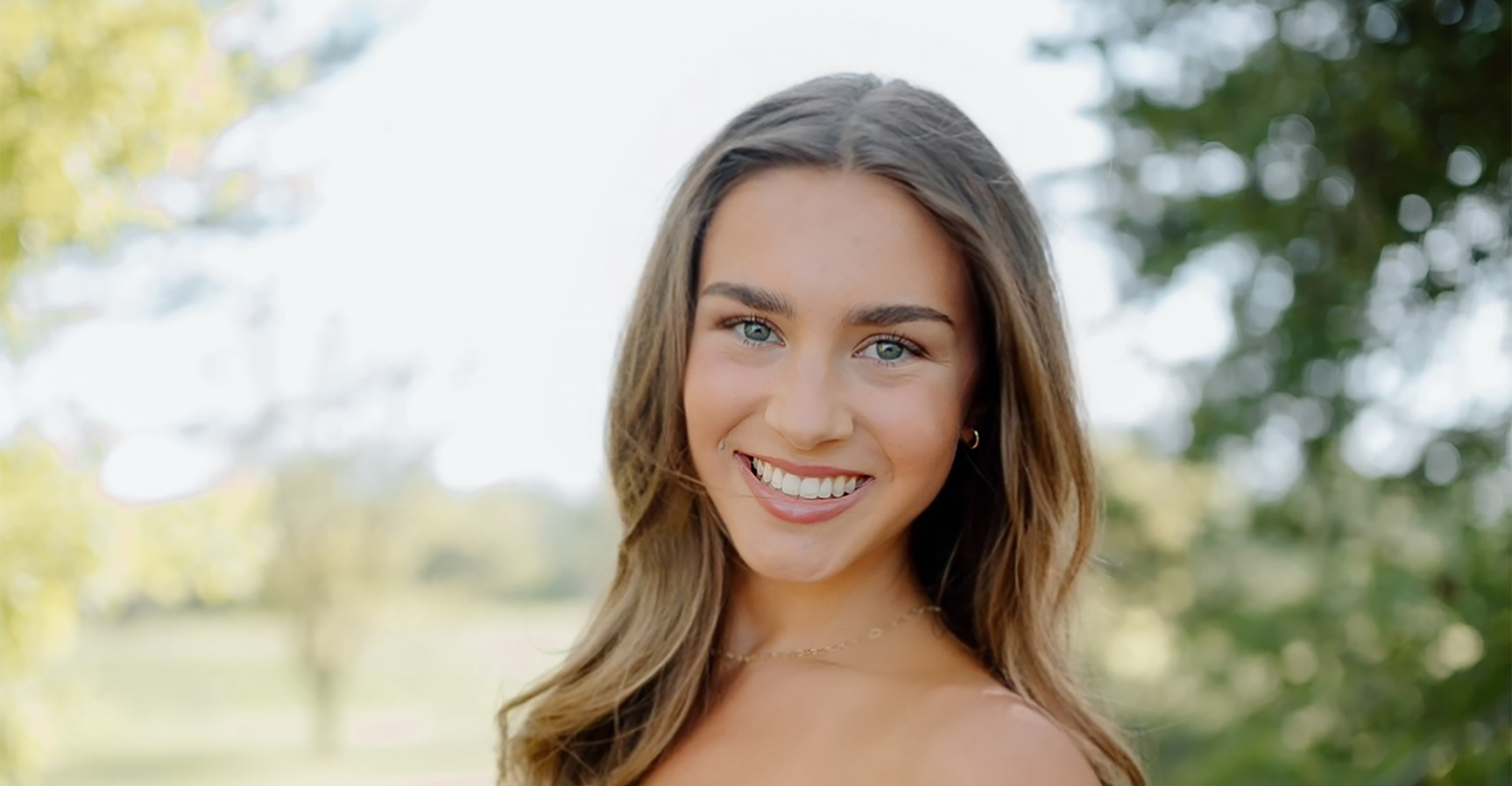 Avery Lay posing in a pink dress in an outdoor setting. Trees are present in the background, but out of focus.