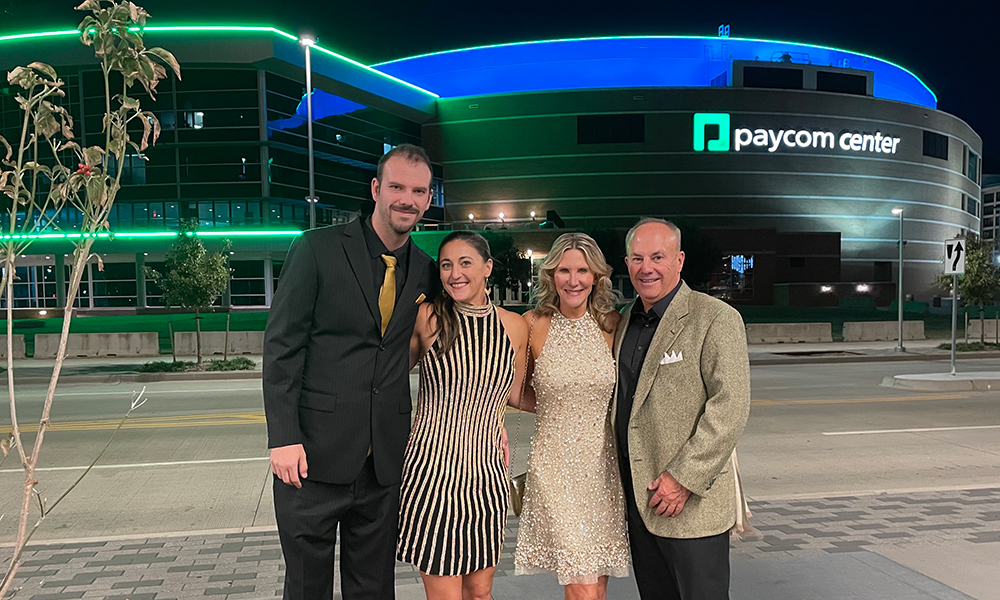 Four adults stand outside Paycom Center as it glows green.