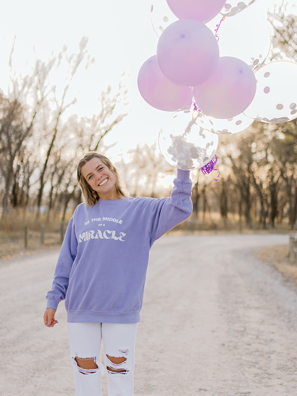 A teenage girl wears a purple sweatshirt and holds purple balloons