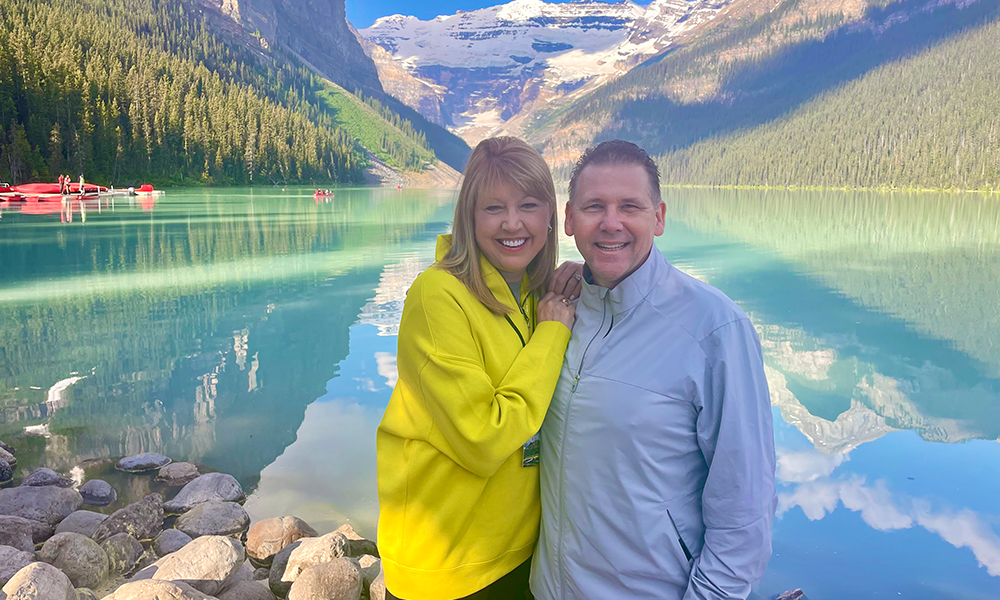 Vickie and Jeff Carr smiling on vacation in front of water and mountains