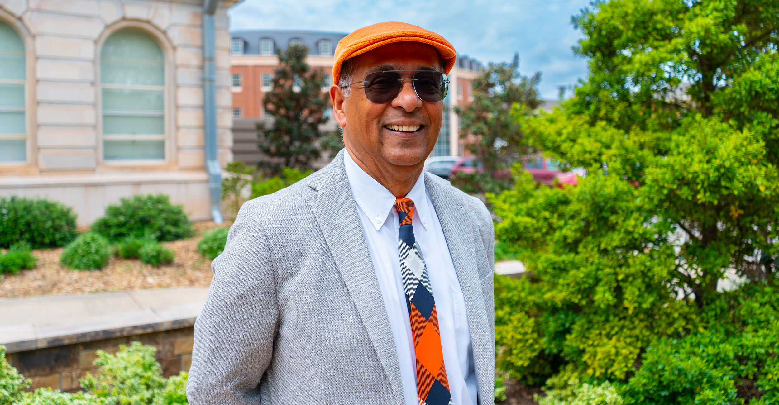 A person wearing an orange flat cap, sunglasses, a light gray blazer, and an orange‑and‑black patterned tie stands outdoors on a university campus walkway, posed for a professional portrait with academic buildings and landscaping in the background.