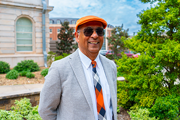 A person wearing an orange flat cap, sunglasses, a light gray blazer, and an orange‑and‑black patterned tie stands outdoors on a university campus walkway, posed for a professional portrait with academic buildings and landscaping in the