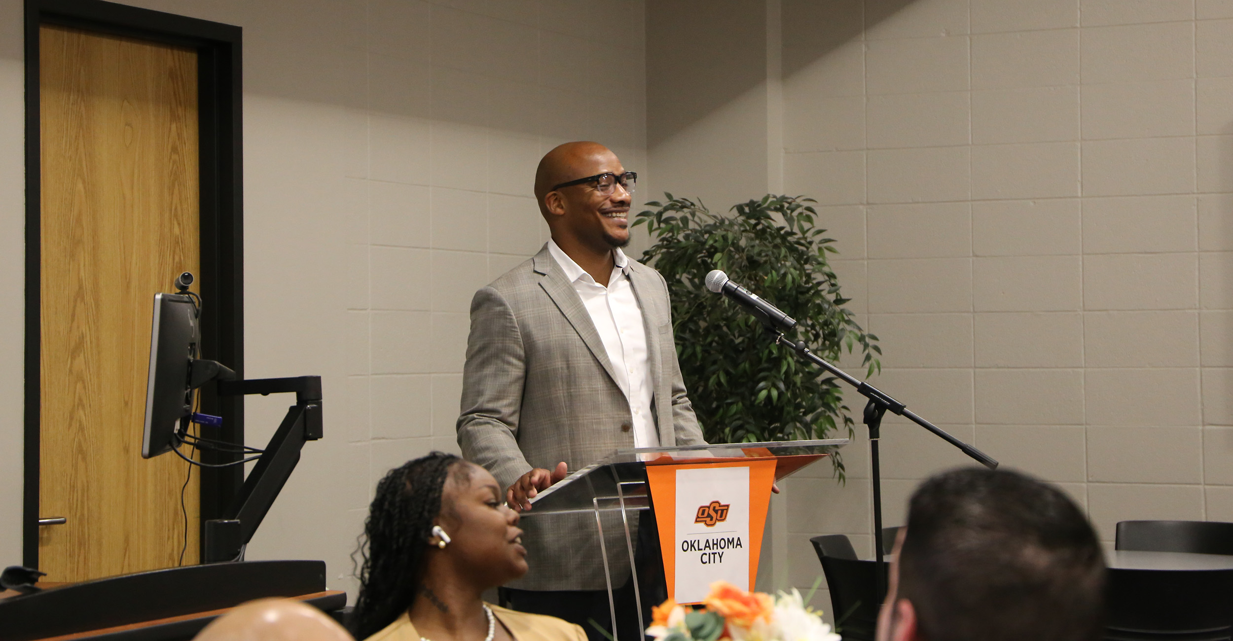 Person standing at a podium with an Oklahoma State University-Oklahoma City logo, speaking into a microphone in a conference room with chairs, a computer station, and a potted plant in the background.