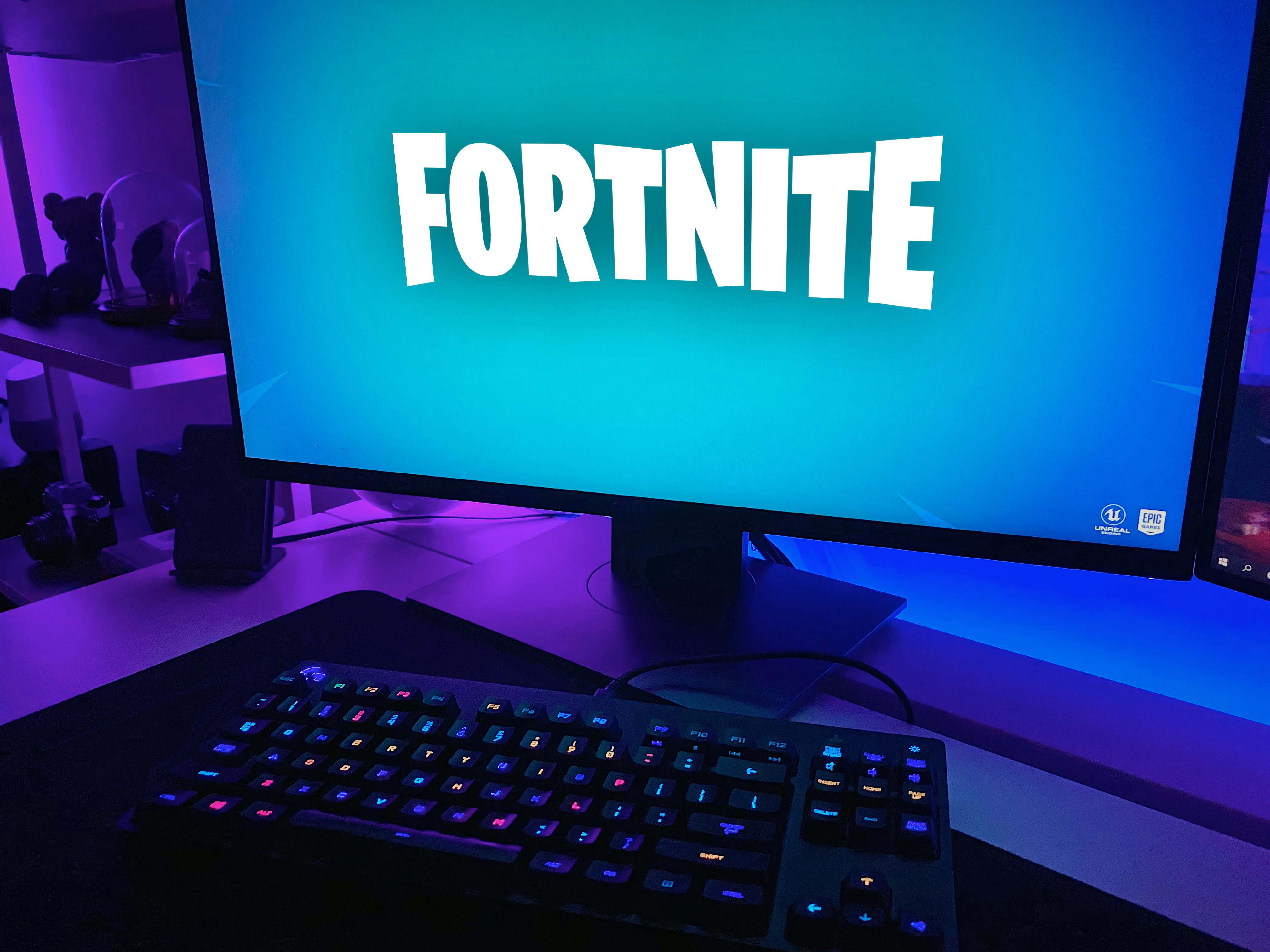 The Fortnite title screen appears on a gaming computer's monitor. A keyboard features glowing keys.