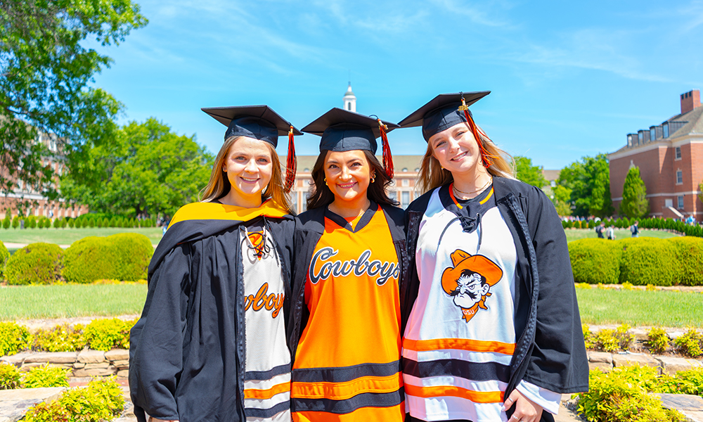Three Oklahoma State University graduates wearing black caps and gowns over OSU hockey jerseys stand together outdoors on campus, posed in front of landscaped greenery and academic buildings for a graduation portrait.